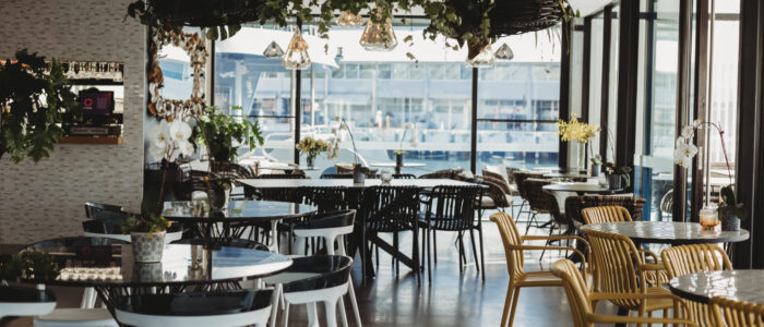 Interior shot of a dinning area. Multiple round tables, greenery hangs on walls, views of the wharf viewable through windows