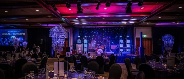 A decorated banquet hall prepared for an event, featuring round tables covered with dark linens, elegant table settings, and tall crystal centerpieces. A stage with colorful lights, a drum kit, and "Awards of Excellence" signage is in the background.