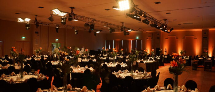 A warmly lit banquet hall set for a formal event, with round tables covered in patterned white tablecloths and surrounded by black chairs. Each table is decorated with tall vases holding orange flowers, along with neatly arranged place settings and folded napkins. Overhead lighting rigs and soft orange wall uplighting complete the ambiance.