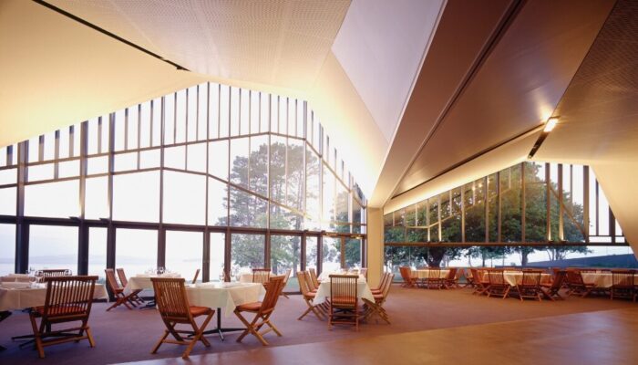 Interior of a modern building with asymmetrical floor to ceiling windows. The floors are an open layout with tables and chairs scattered throughout. The chairs are an orange wooden chairs with the tables set with a white tablecloth and wine glasses. The large windows let in lots of natural light illuminating the room.