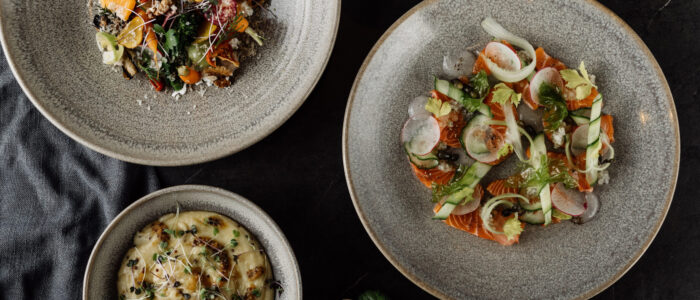 An artistic photograph of professionally created meals viewed from above. Meals sit in stone plates upon a black table cloth