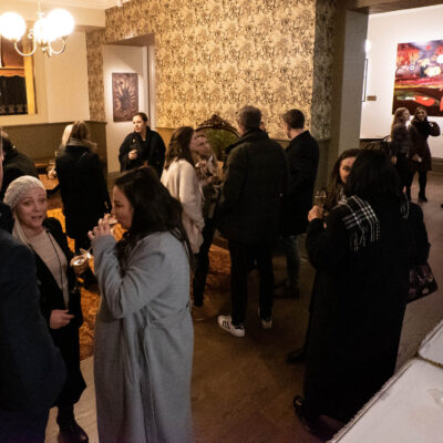 A dimly lit lounge with a vintage aesthetic, where guests in winter clothing are mingling, holding drinks, and engaging in conversation. A table with bottles and glasses is visible in the foreground, while a warmly lit adjoining room with red decor can be seen in the background.