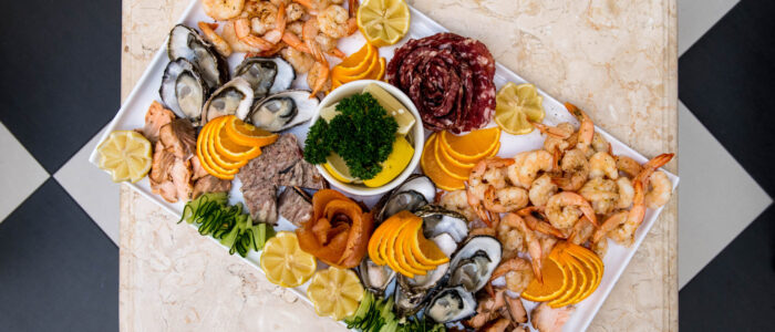 A top-down view of a vibrant seafood platter filled with shrimp, oysters, salmon, sliced citrus, and cured meats, beautifully arranged on a white tray.