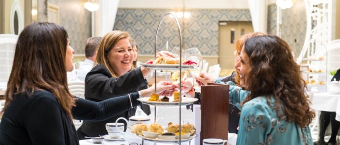 A group of four women enjoys an elegant afternoon tea in a bright, ornate tearoom, raising their glasses in a toast over a three-tiered tray of pastries and scones.