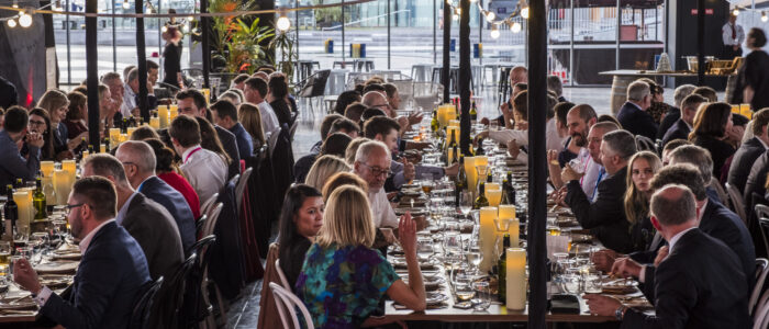 room filled with people in business attire sitting at long tables. Large wall of windows in the background looking out to the street