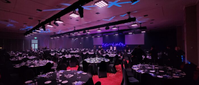 An event space illuminated by soft purple and blue lights, showcasing tables adorned sparkling glassware. A truss with lighting equipment is suspended overhead. Staff are busy around the room setting up the event space