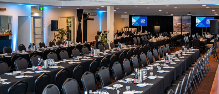 A banquet hall with long tables draped in black tablecloths, set with plates, glassware, and cutlery. Rows of black chairs surround the tables. The back wall has to screens displaying "2024 Annual Service Awards". The ceiling is adorned with string lights.