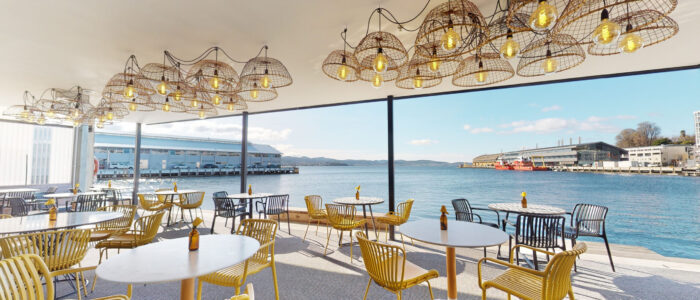 Large room with large windows over looking the wharf, very well lit with multiple round tables and yellow chairs