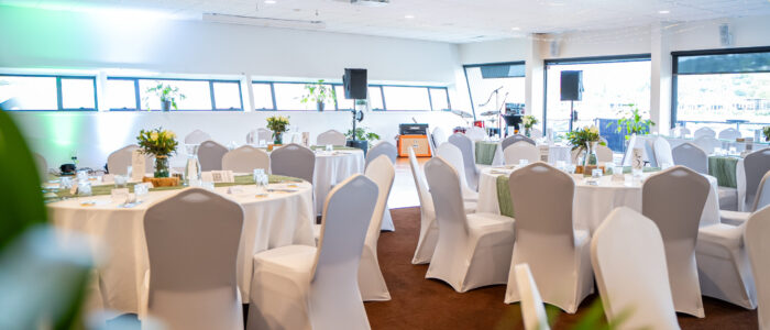 A decorated event space with round tables covered in white tablecloths and green table runners. The tables are elegantly set with glassware, cutlery, and floral centerpieces. White chairs with covers surround the tables, and a small stage with musical instruments is visible in the background.