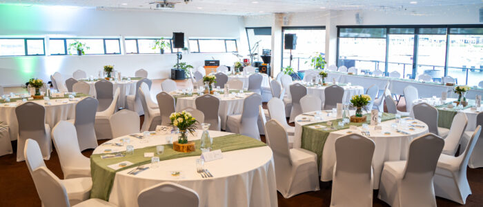 A decorated event space with round tables covered in white tablecloths and green table runners. The tables are elegantly set with glassware, cutlery, and floral centerpieces. White chairs with covers surround the tables, and a small stage with musical instruments is visible in the background.