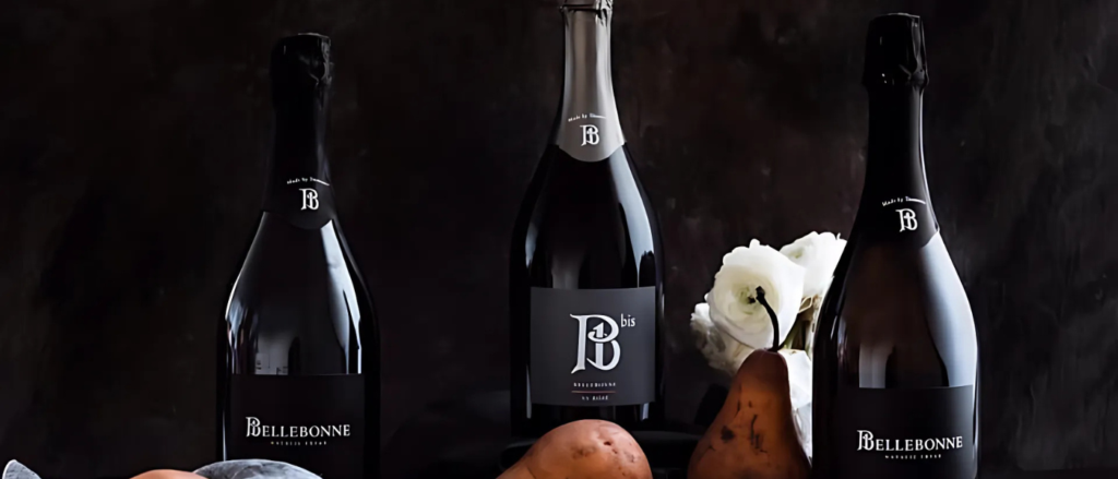 The image shows three bottles of Bellebonne sparkling wine, a plate with a wedge of cheese and a knife, two pears, and some white flowers. The background is dark, creating a contrast with the lighter elements in the foreground.