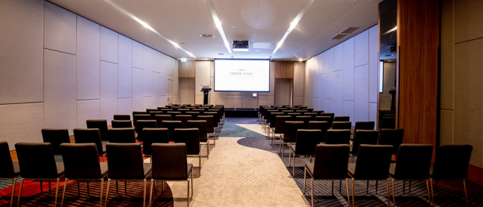 An empty conference room with rows of black chairs arranged facing a podium and a large screen at the front. The room has a modern design with light-colored walls, carpeted flooring, and ceiling lights.