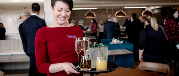 A person in a red dress holding a tray with various drinks, including two glasses of champagne, a glass of what appears to be an alcoholic beverage with ice and mint leaves, another glass with an amber-colored liquid, and a glass of orange juice. In the background, there are several people engaged in conversation in what appears to be a social or professional gathering space.
