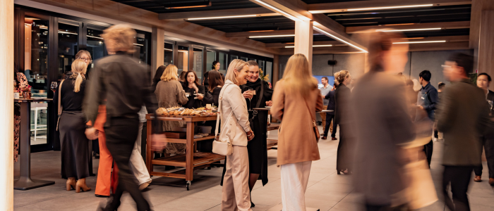 A group of people in semi-formal attire socializing in a modern indoor space with large windows and overhead lighting. Tables with food and drinks are visible, indicating a professional or business-related event.