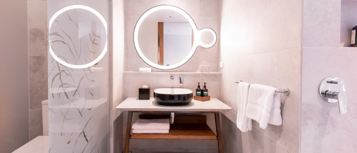 A modern bathroom with a large, illuminated oval mirror above a sink. The sink is black and sits on a white countertop with wooden shelving underneath, holding neatly folded towels. To the left of the sink area is a glass shower door with an abstract design. On the right side of the image, there is a towel rack holding a white towel and part of a chrome faucet handle is visible. The bathroom has light-colored tiles on the walls and floor, creating an overall clean and contemporary look.