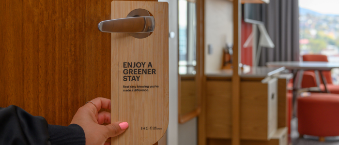 A hand holding a door handle with a wooden sign hanging from it. The sign reads: "ENJOY A GREENER STAY Rest easy knowing you've made a difference." The bottom of the sign includes the IHG Hotels & Resorts logo and text.