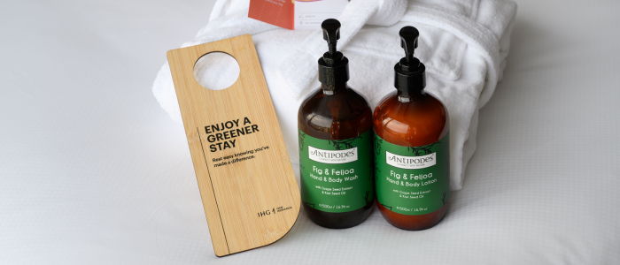 Two bottles of body wash labeled "Fig & Feijoa Hand & Body Wash" from the brand "Antipodes," placed on a white bathrobe. There is also a wooden door hanger with the text "ENJOY A GREENER STAY" and the logo "IHG Hotels & Resorts."