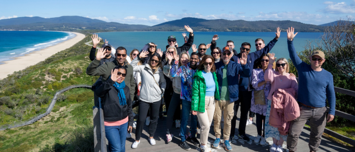A group of people is standing on a wooden platform overlooking a scenic coastal landscape. The background features a long sandy beach, blue ocean waters, and distant hills under a partly cloudy sky. The individuals are waving at the camera.