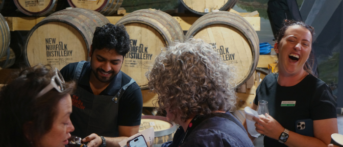 The image shows a group of people standing in front of several wooden barrels labeled 'NEW NORFOLK DISTILLERY.' The people appear to be engaged in a tasting or tour, as one person is holding a glass, and another is holding a bottle.