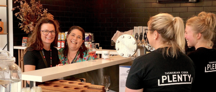 The image shows a group of people in a bar or café setting. There are two people behind the counter, two of whom are wearing black shirts with the text 'TASMANIAN CIDER PLENTY' on the back. They appear to be serving or preparing drinks. In front of them, there are two customers standing at the counter. The background includes various items such as bottles, a coffee machine, and some decorative elements like dried plants.