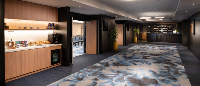 A modern, well-lit hallway or lobby area with a patterned blue and white carpet. On the left side, there is a wooden counter with various items including snacks, beverages, and a coffee machine. There is also a small refrigerator under the counter. In the background, there are doors leading to other rooms and some potted plants placed along the walls. On the right side of the image, there is another counter area that appears to be set up as a bar or refreshment station with bottles and glasses displayed on shelves behind it. The ceiling has circular light fixtures that add to the contemporary design of the space.
