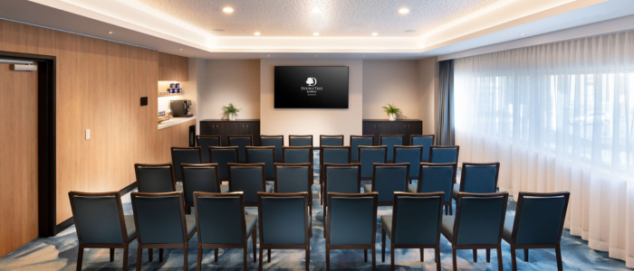 A small conference room or meeting space with rows of black chairs facing a large flat-screen television mounted on the wall. The room has wooden paneling on the left side, a shelf with some items, and large windows covered with sheer curtains on the right side. The floor is carpeted with a blue and white pattern. There are recessed lights in the ceiling providing illumination. The television screen displays the logo of 'DoubleTree by Hilton.'