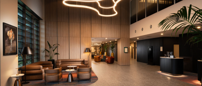 A modern hotel lobby with a spacious and stylish design located at DoubleTree by Hilton Hobart. The lobby features wooden paneling on the walls, a large abstract light fixture hanging from the ceiling, and contemporary furniture including brown sofas and armchairs. There is a reception desk on the right side of the image, and large potted plants add greenery to the space. A painting of flowers hangs on the left wall, and there are several tables with lamps placed around the seating area.