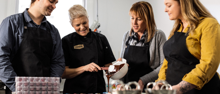 Four people standing around a table, engaged in a cooking or baking activity. They are all wearing aprons, and one person is pouring chocolate from a bowl while another person holds the bowl. Various kitchen utensils and containers are on the table in front of them.