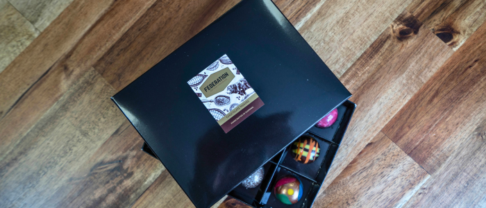 An open black box of assorted chocolates on a wooden surface. The lid of the box has a label with the text 'Federation' and images of cocoa beans. Inside the box, there are various colourful chocolates arranged in individual compartments.