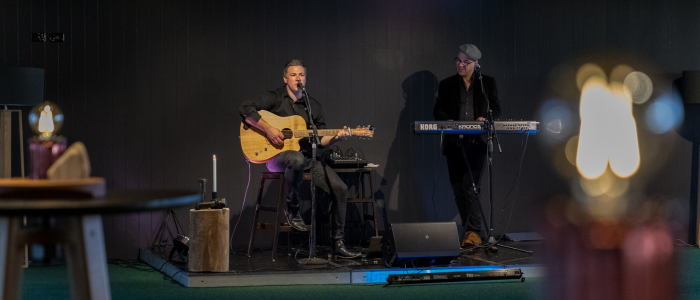 2 people standing on stage, one holding a guitar singing the other looking towards him as he plays the keyboard