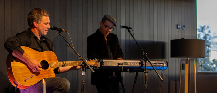 2 people on stage. One playing guitar and the other playing the keyboard. Black wooden wall behind them