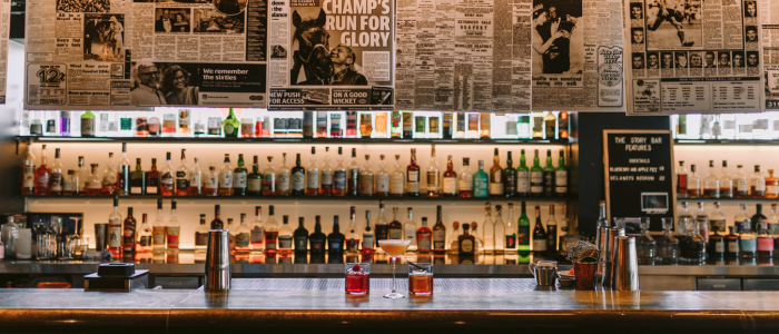 Well-stocked bar with liquor bottles on shelves, two drinks on the counter, a cocktail shaker, and bar tools. Newspaper clippings and headlines are displayed above the bar, creating a vintage ambiance.