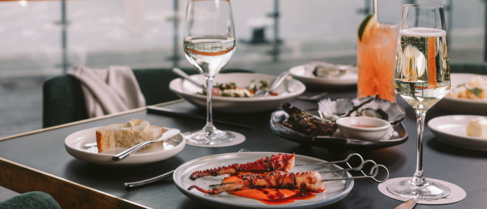 Elegant dining table with plates of grilled octopus skewers in red sauce, oysters or shellfish, and bread slices. Drinks include two glasses of white wine and an orange cocktail with a lemon slice. The background features large windows with a scenic, out-of-focus view.