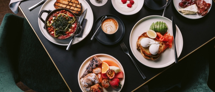 Table set with a variety of breakfast dishes including shakshuka with toast, poached eggs with avocado and smoked salmon, assorted pastries with fruit, a bowl of raspberries, a cheese and cured meat platter, and a cup of coffee with latte art.