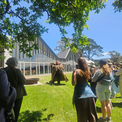 Group of people gathered outdoors on a grassy area near a modern glass building, watching a Welcome to Country Ceremony under a sunny blue sky with trees in the background.