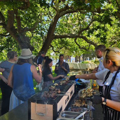 Group of people gathered around an outdoor barbecue under large leafy trees, with a chef grilling skewers on a stainless steel grill and tables set up in the background.