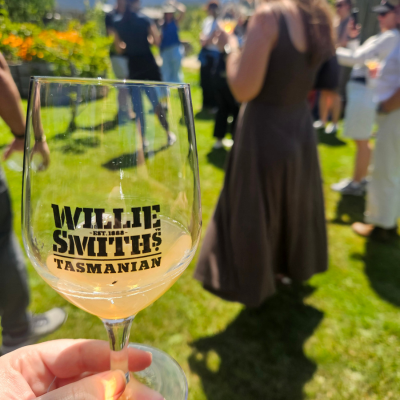 Close-up of a glass with ‘Willie Smith’s Tasmanian’ printed on it, held in the foreground with a light orange drink inside, while people stand on a grassy area in the background during an outdoor event.