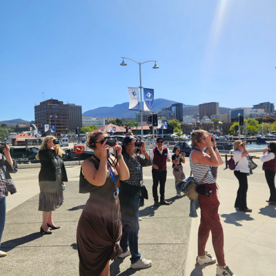 Group of people standing on a waterfront promenade taking photos, with boats docked in the harbor, city buildings, and mountains in the background under a clear blue sky.