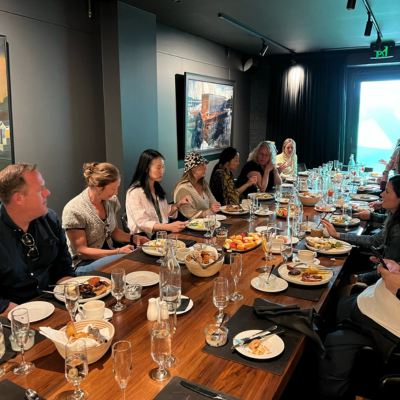 Group of people seated around a long wooden dining table in a modern, dimly lit restaurant, with plates of food, bread baskets, and glasses arranged for a shared meal.