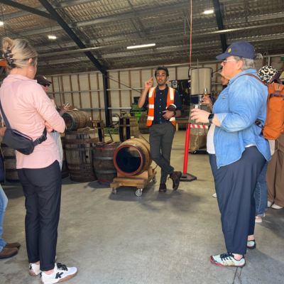 Group of people inside a warehouse-style building listening to a guide wearing a high-visibility vest, with wooden barrels and industrial equipment visible in the background.