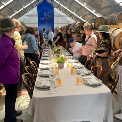 Group of people gathered around a long dining table set with white tablecloth, plates, glasses, and orange napkins inside a barrel room with wooden chairs and rows of large wooden barrels along the walls.