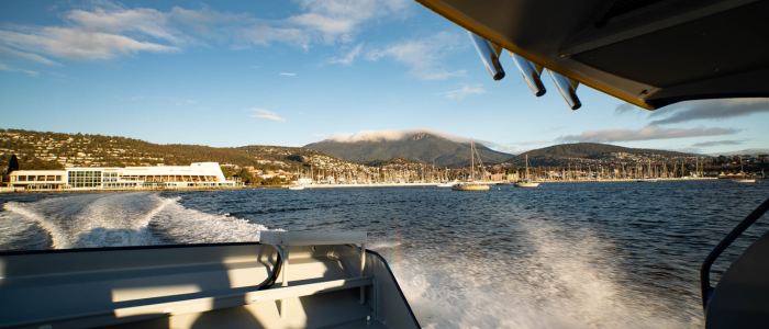 View from the back of a boat showing its wake trailing across calm blue water, with a marina full of sailboats and a coastal town in the background, and a mountain partially covered by clouds under a clear sky.
