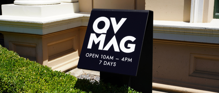 A black metal sign that reads "QVMAG. Open 10am - 4pm, 7 Days" This sits hanging over a green, neatly trimmed hedge with a stone building behind