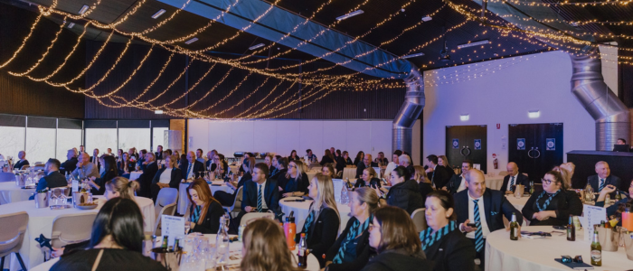 A large banquet hall filled with attendees dressed in formal attire, seated at round tables. The ceiling is adorned with warm string lights, creating an elegant atmosphere. Many guests appear engaged, looking toward an unseen speaker or presentation.