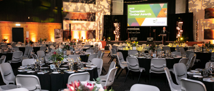 A beautifully decorated event hall set up for the 2019 Tasmanian Timber Awards. Round tables with black tablecloths, fine glassware, and floral centerpieces fill the room. A stage with a large screen displaying the event name is in the background, along with warm ambient lighting.