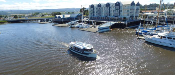 A small passenger boat moves through shimmering waters near a marina, with waterfront buildings and docked sailboats in the background.