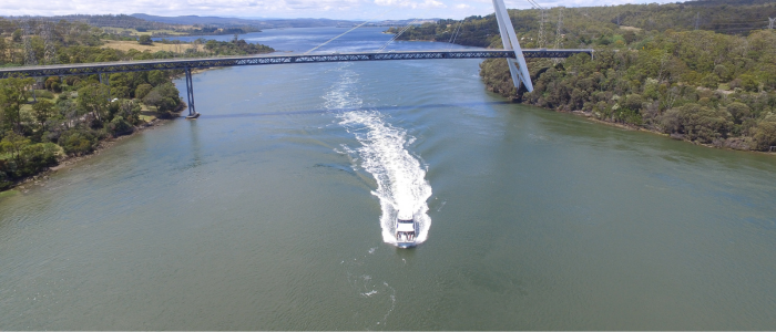 A boat speeds along a wide river, leaving a foamy trail behind as it passes under a sleek suspension bridge, surrounded by forested hills.
